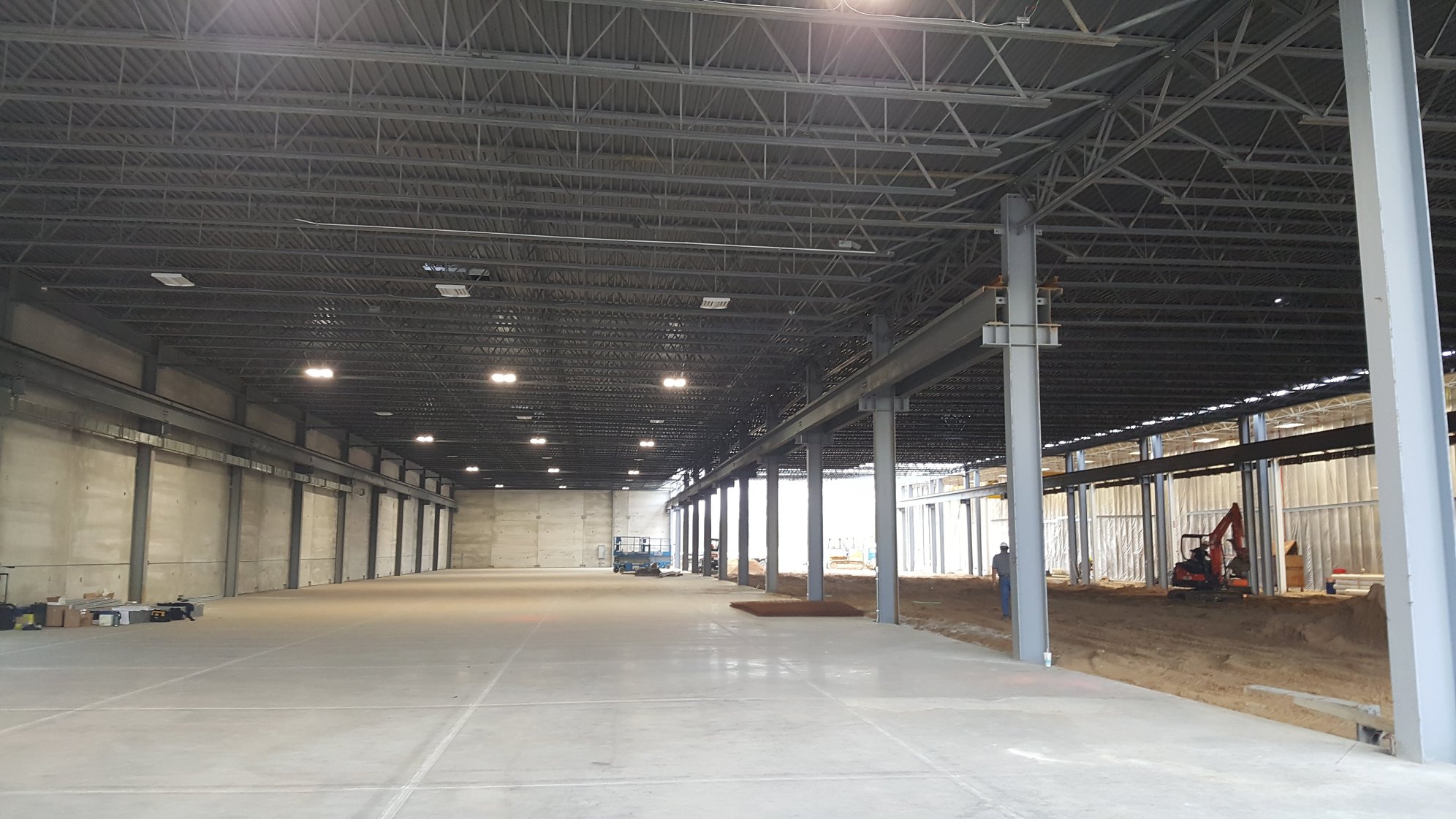 Interior view of a large industrial warehouse under construction with steel beams, concrete floor on the left, and exposed soil with a small excavator and workers on the right side.